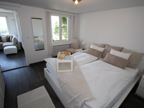 A modern bedroom with a large bed, bedside tables, a nightstand, a mirror, and a view of a living area. - Kitty (Parterre) Appartement/Fewo, Dusche oder Bad, WC 4-Bettwohnung by Interhome (Gstaad)