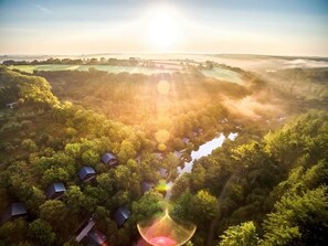 A rural landscape with houses surrounded by dense forest, a river, and a sunrise.