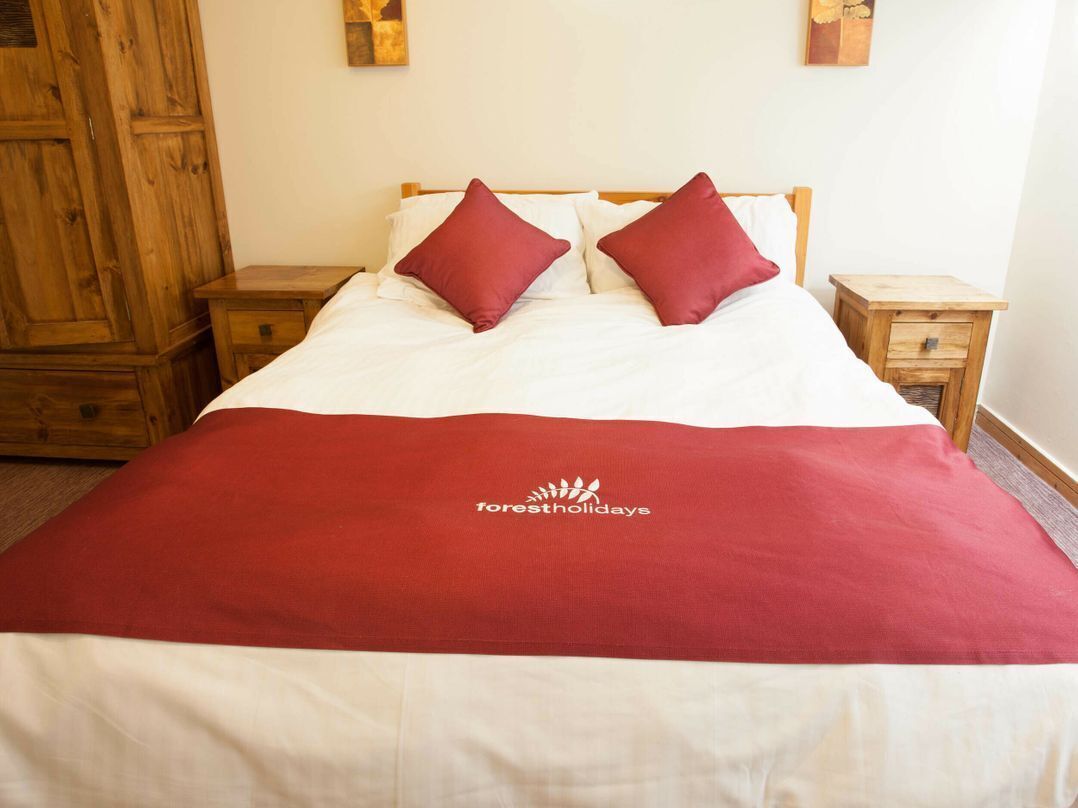 Een bed met een rode deken en twee kussens, een houten kledingkast en een klein nachtkastje met een lade.