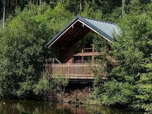 Mökki, jossa on puinen terassi, ja sen ympärillä on tiheää metsää sekä vesistö.
