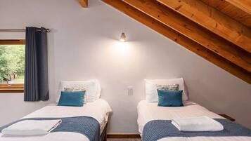 A bedroom with two beds, wooden ceiling beams, a window with curtains, and a wall-mounted light.