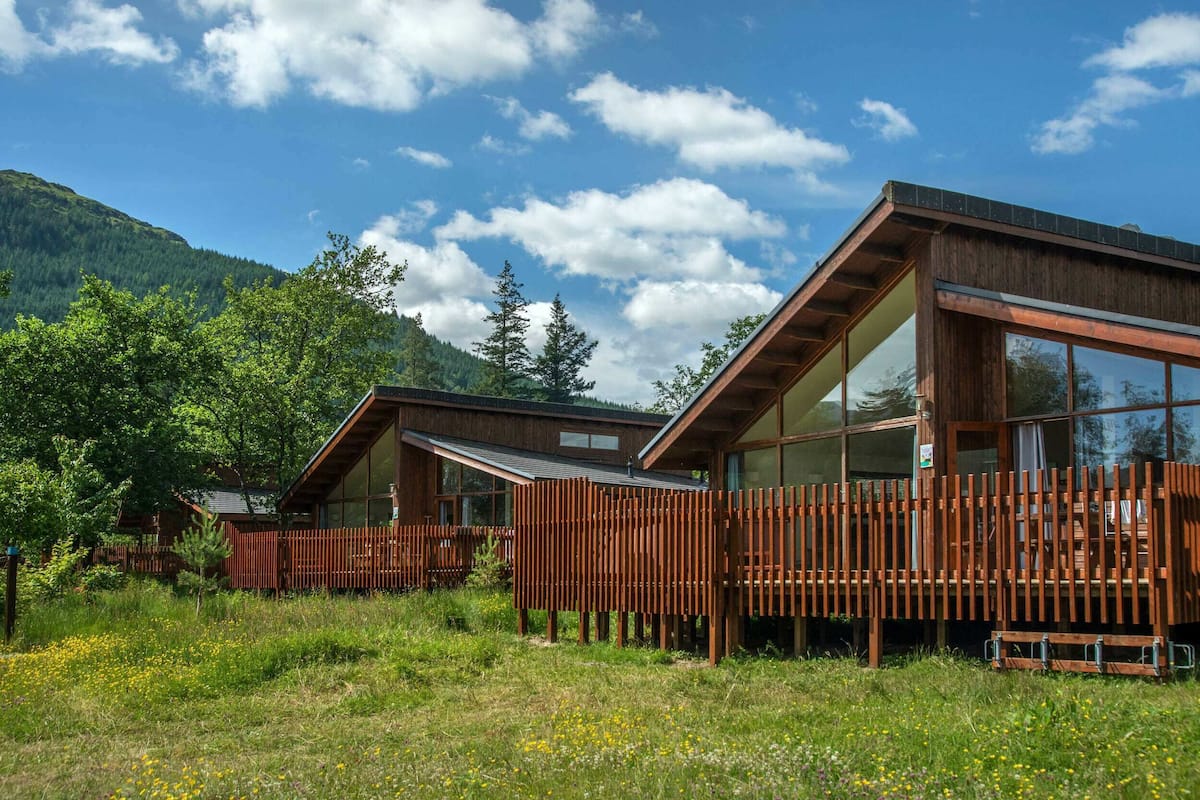 Una cabaña moderna de madera con ventanas amplias y una terraza de madera, rodeada de abundante vegetación y montañas.