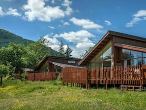 Una cabaña moderna de madera con ventanas amplias y una terraza de madera, rodeada de abundante vegetación y montañas.