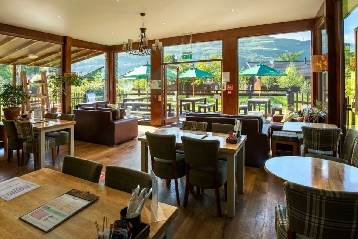 A restaurant with wooden tables and chairs, large windows with views of greenery and hills, and a cozy seating area.