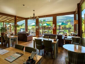 A restaurant with wooden tables and chairs, large windows with views of greenery and hills, and a cozy seating area. - Ardgartan Argyll - Forest Holidays (Arrochar)