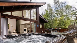 A hot tub area with a wooden deck, wicker chairs, and a view of a modern house with large windows.