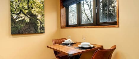 A dining area with a table set for two, a painting of trees, and a window with a view of trees.