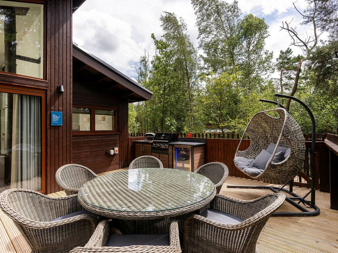 A wooden deck with a glass-top table surrounded by wicker chairs, a hanging chair, and a barbecue grill.