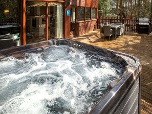 A hot tub on a wooden deck with outdoor furniture, a grill, and a glass-enclosed building.