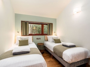 A room with two beds, a window with curtains, a small round table, and a wall-mounted light fixture. - Glentress Forest - Forest Holidays (Peebles)