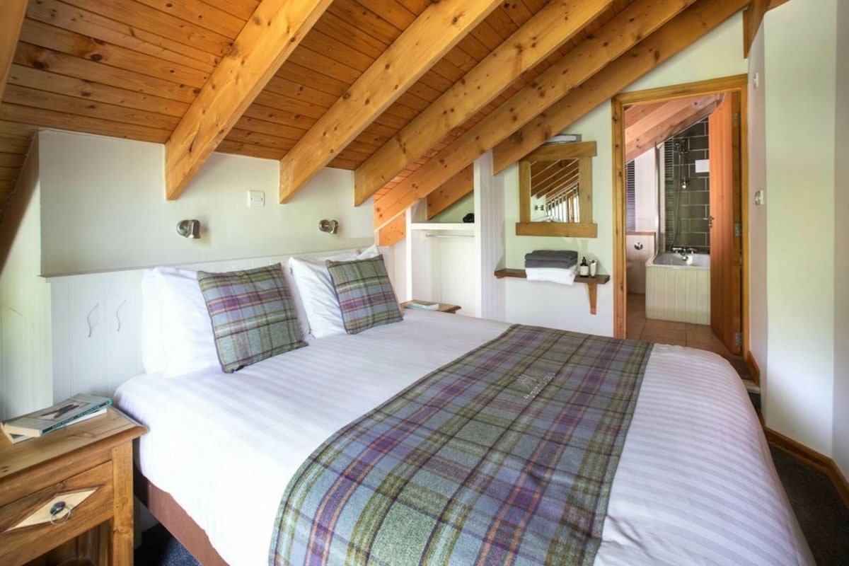 A bedroom with a wooden ceiling, a large bed with a plaid blanket, a nightstand, a mirror, and a door leading to a bathroom.
