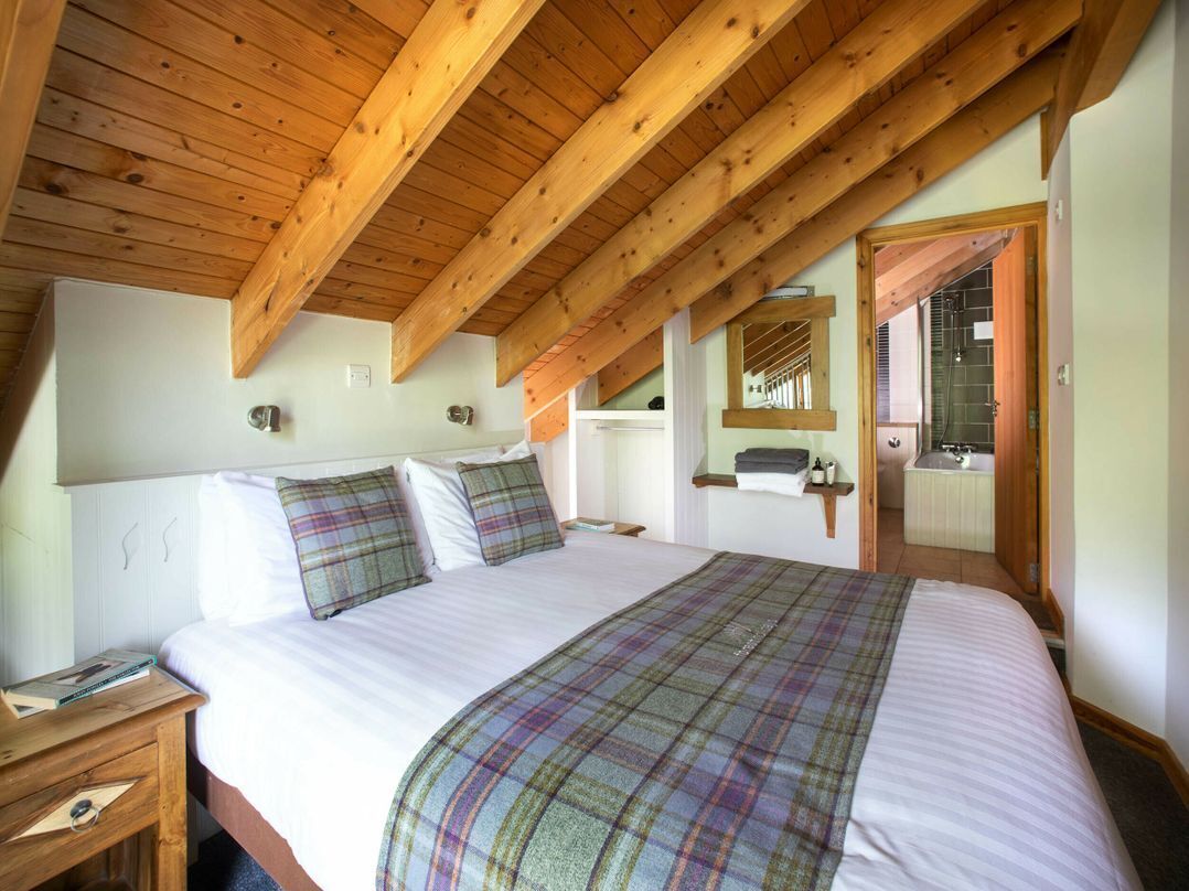 A bedroom with a wooden ceiling, a large bed with a plaid blanket, a nightstand, a mirror, and a door leading to a bathroom.