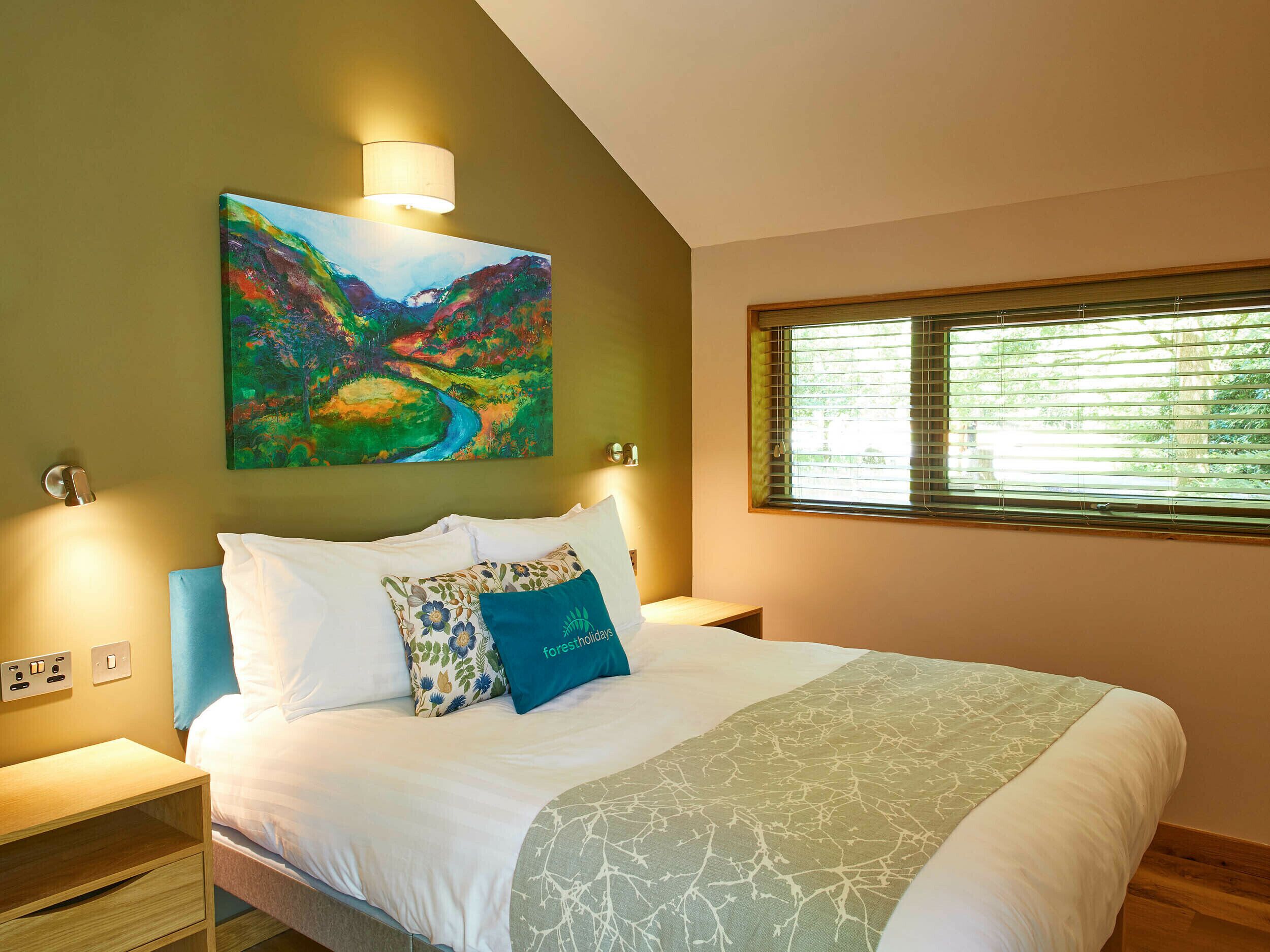 Una camera da letto con un letto, cuscini, un comodino, una lampada a muro, una finestra con persiane e un quadro appeso al muro.