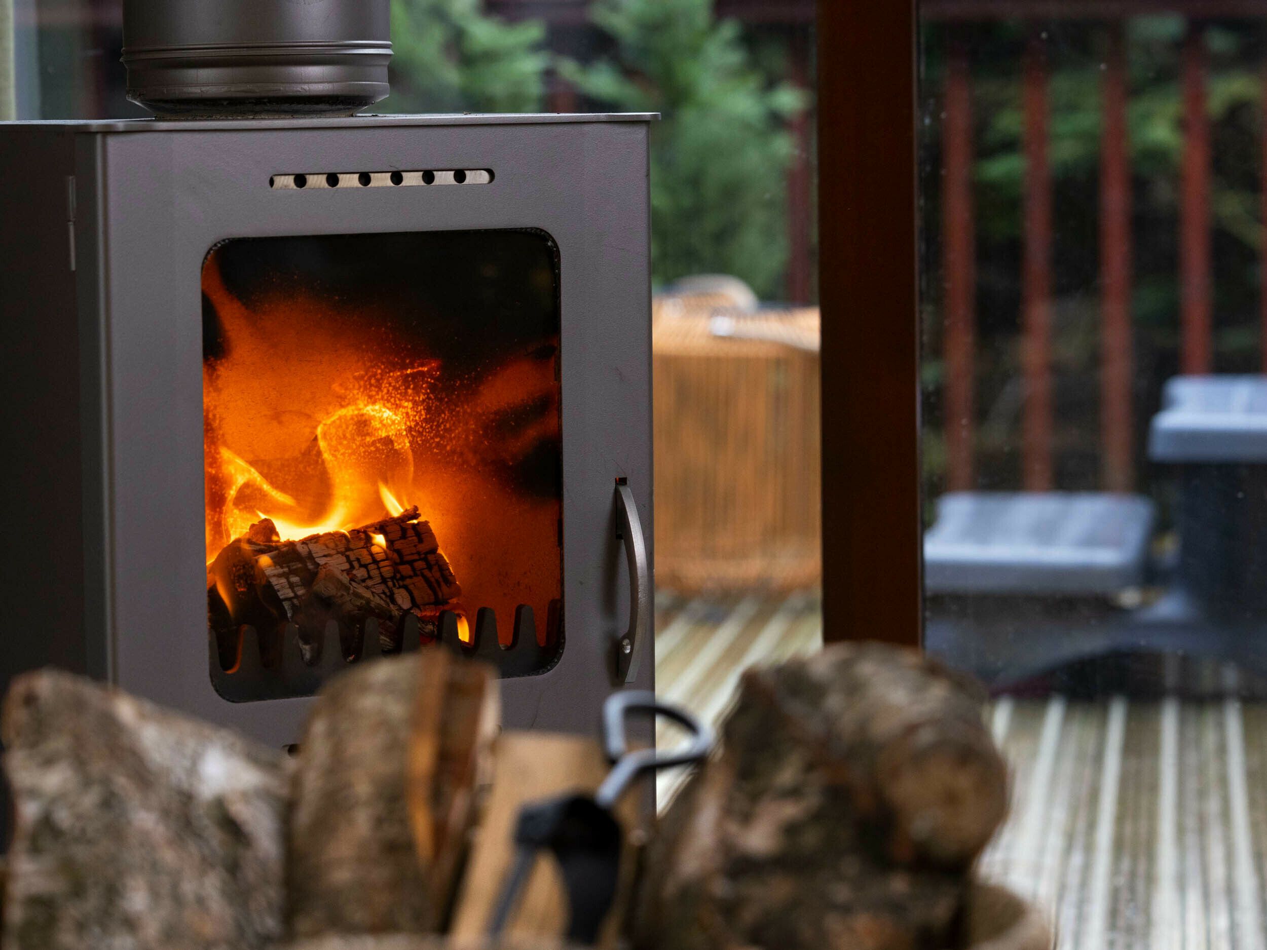 A wood-burning stove with visible flames and logs inside, placed on a wooden deck.