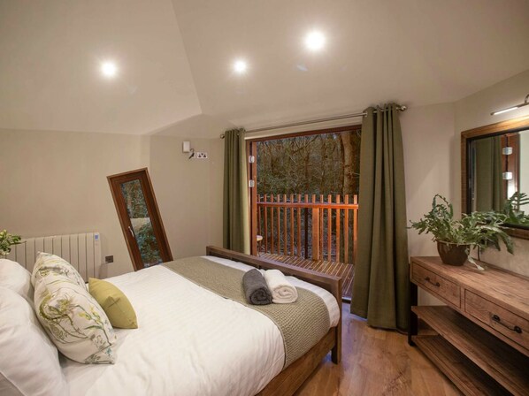A bedroom with a bed, a wooden dresser, a mirror, and a window with a view of trees. - Garwnant Brecon Beacons- Forest Holidays (Merthyr Tydfil)