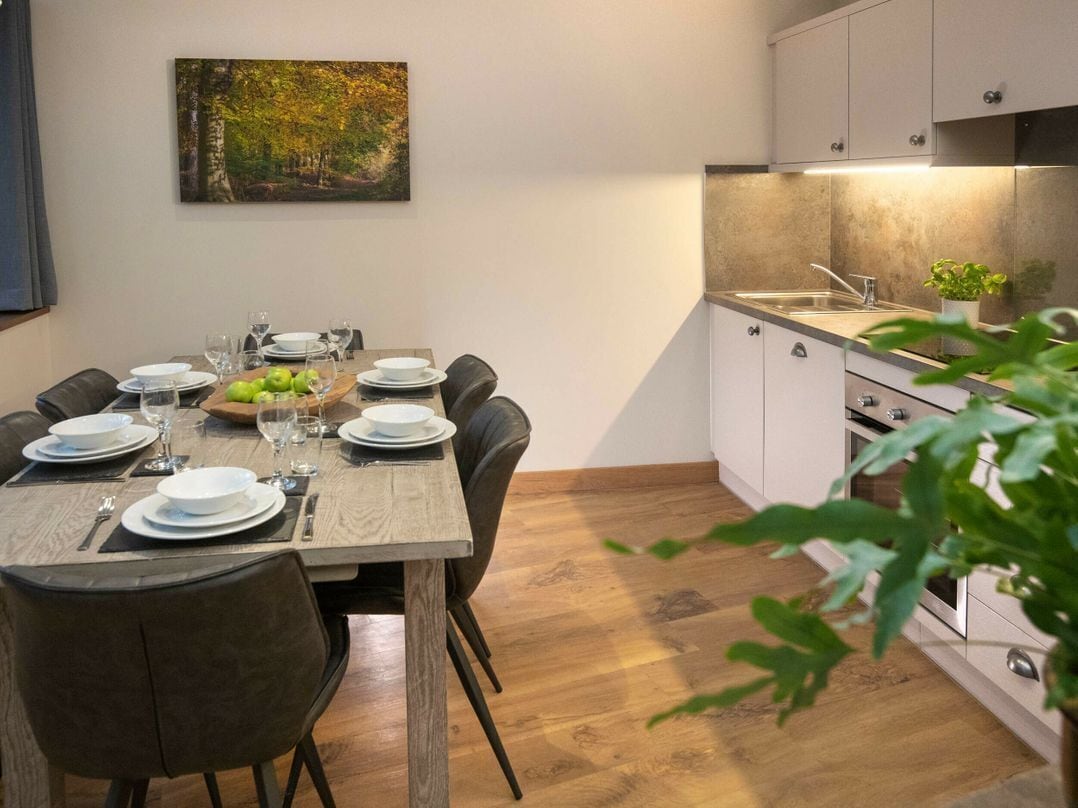 A dining area with a wooden table set for four, a painting on the wall, and a kitchen with white cabinets and a countertop.