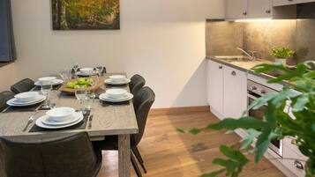 A dining area with a wooden table set for four, a painting on the wall, and a kitchen with white cabinets and a countertop.