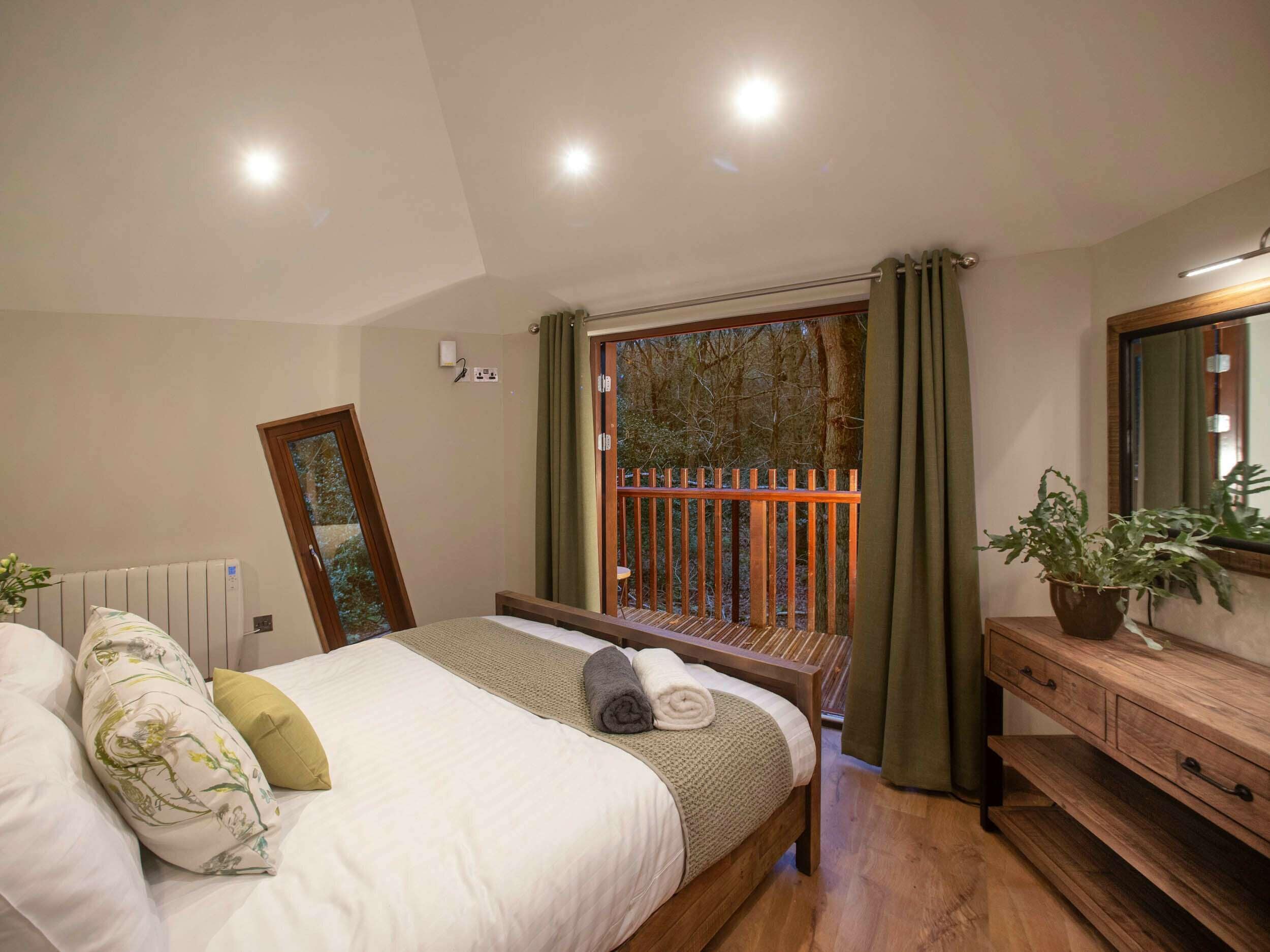 A bedroom with a bed, a wooden bench, a dresser, a mirror, and a view of a balcony with a railing.