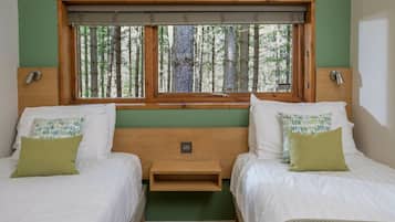 A room with two beds, a window with a view of trees, a wooden headboard, and a small bedside table.