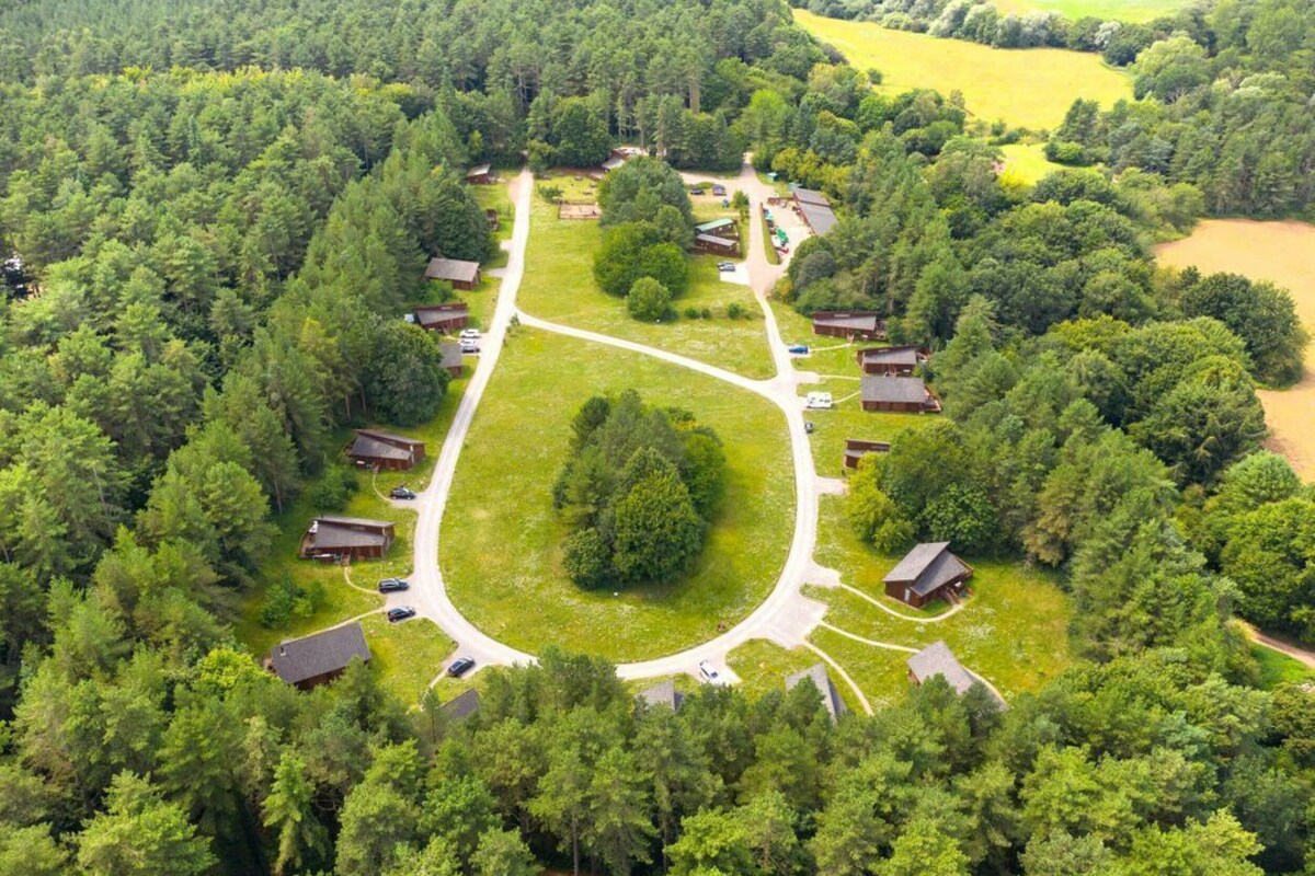 Vue aérienne d’un terrain de camping comprenant plusieurs cabanes, une grande pelouse centrale et une forêt dense qui l’entoure.