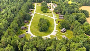 Vue aérienne d’un terrain de camping comprenant plusieurs cabanes, une grande pelouse centrale et une forêt dense qui l’entoure.