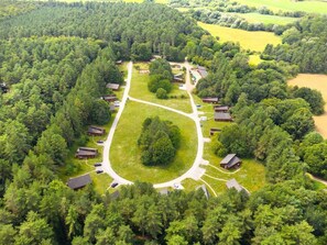 Vue aérienne d’un terrain de camping comprenant plusieurs cabanes, une grande pelouse centrale et une forêt dense qui l’entoure.