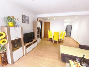 A modern living room with a TV, dining area with yellow chairs, and a kitchen in the background. - Apartment Sandra Crikvenica (Crikvenica)