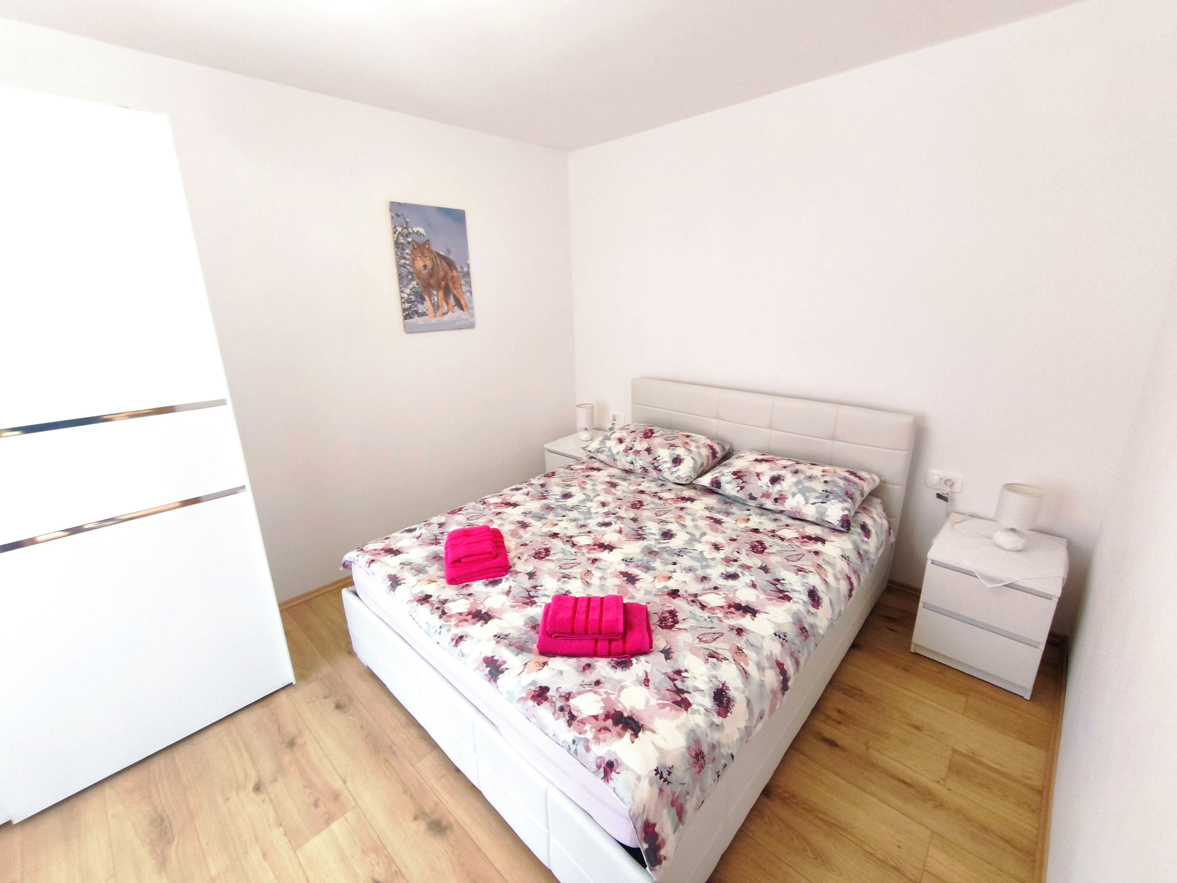 Un dormitorio con una cama, una mesita de noche, una lámpara, un cuadro en la pared y suelo de madera.