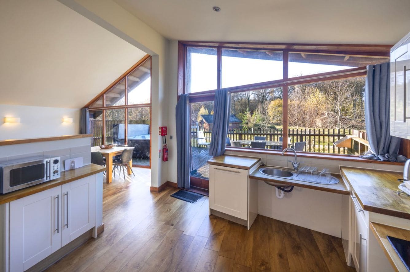 A modern kitchen with wooden flooring, a dining area with a table and chairs, and large windows offering a view of a fenced outdoor area.