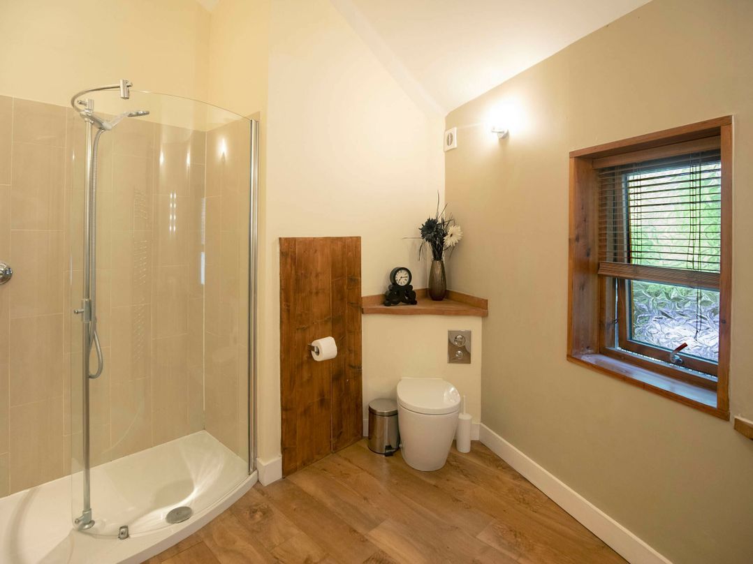 Un baño con cabina de ducha de vidrio, inodoro, un pequeño cubo de basura, un estante de madera con un reloj y flores, y una ventana con persianas.