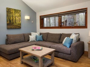 Uma sala de estar com sofá seccional, mesa de centro, pintura de uma floresta, janela com vista para árvores e luminária fixada na parede.