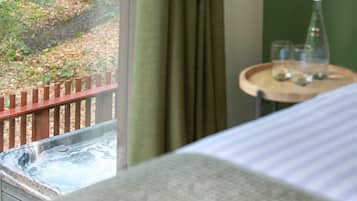 A hotel room with a view of a wooden deck and greenery outside. There is a small round table with a bottle and glasses on it.