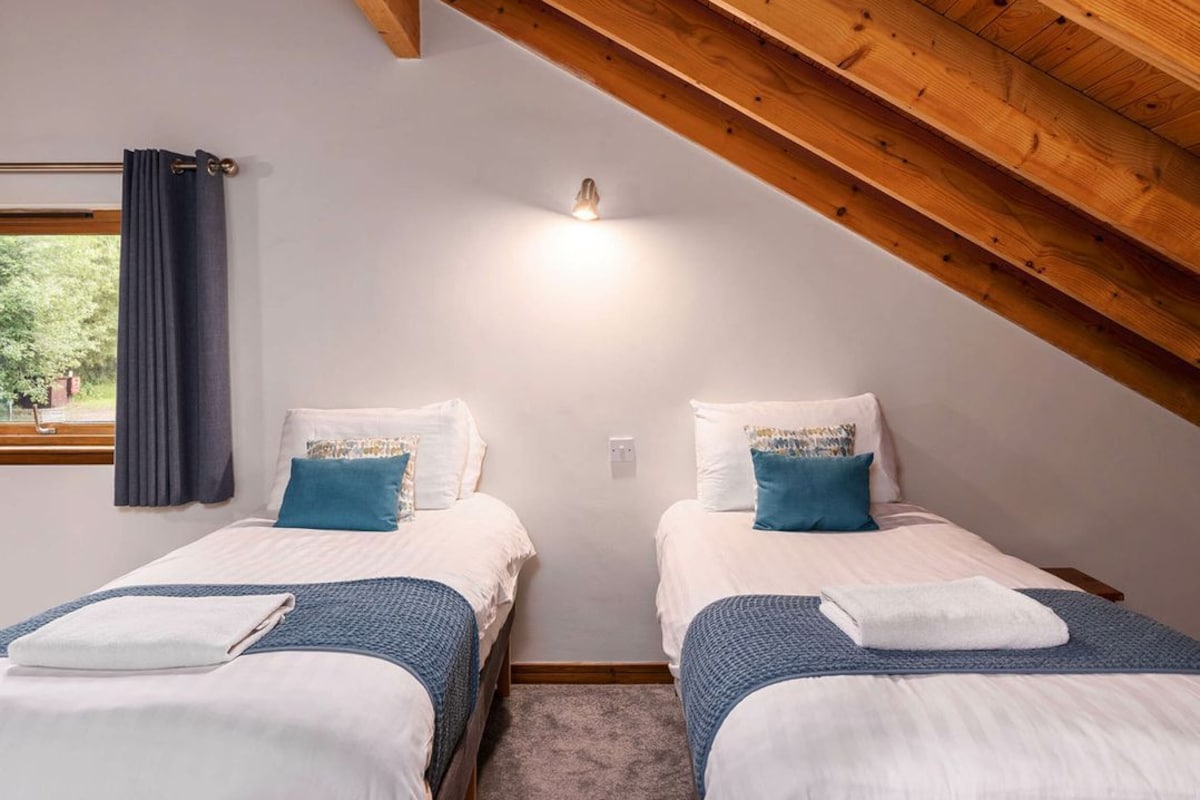 A bedroom with two beds, a window with curtains, a wooden ceiling, and a wall-mounted light fixture.