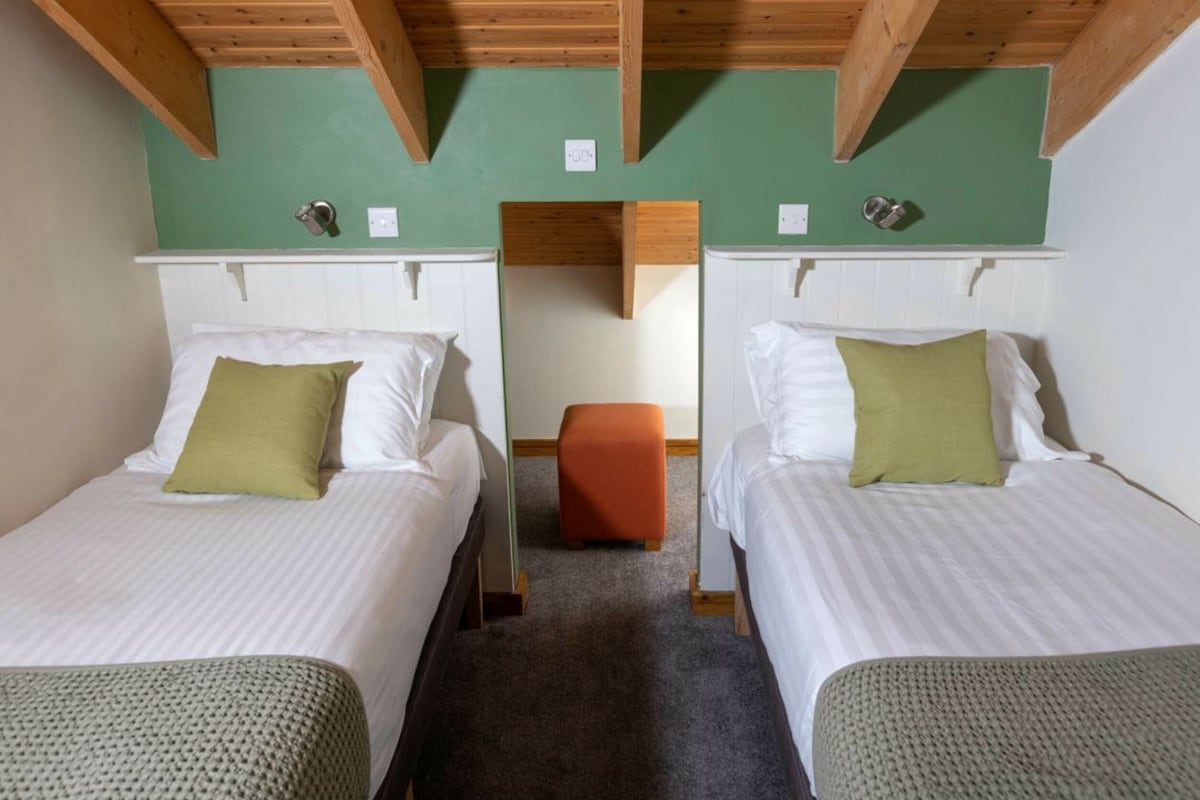 A room with two beds, a small orange ottoman, and wooden ceiling beams.