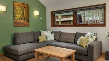 A living room with a sectional sofa, a wooden coffee table, a painting of a forest, and a window with a view of trees.