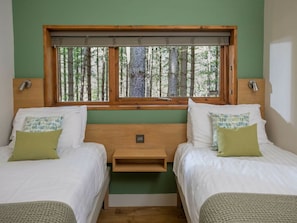 Una habitación con dos camas, una ventana que da a árboles, cabecero de madera y una mesita de noche pequeña.