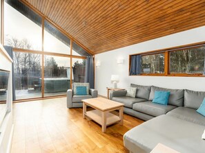 A modern living room with a wooden ceiling, large windows, a grey sofa, a wooden coffee table, and a small side table. - Cropton Forest - Forest Holidays (Pickering)