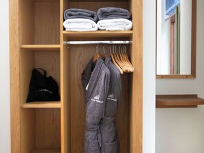 Um guarda-roupa de madeira com toalhas dobradas, uma mala preta e roupões cinzentos pendurados.