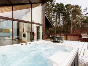 A hot tub on a wooden deck with a view of a modern house and forest.