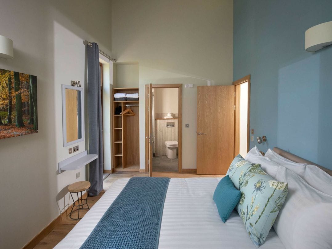 A bedroom with a large bed, a bedside table, a wardrobe, a bathroom visible through an open door, and a painting on the wall.