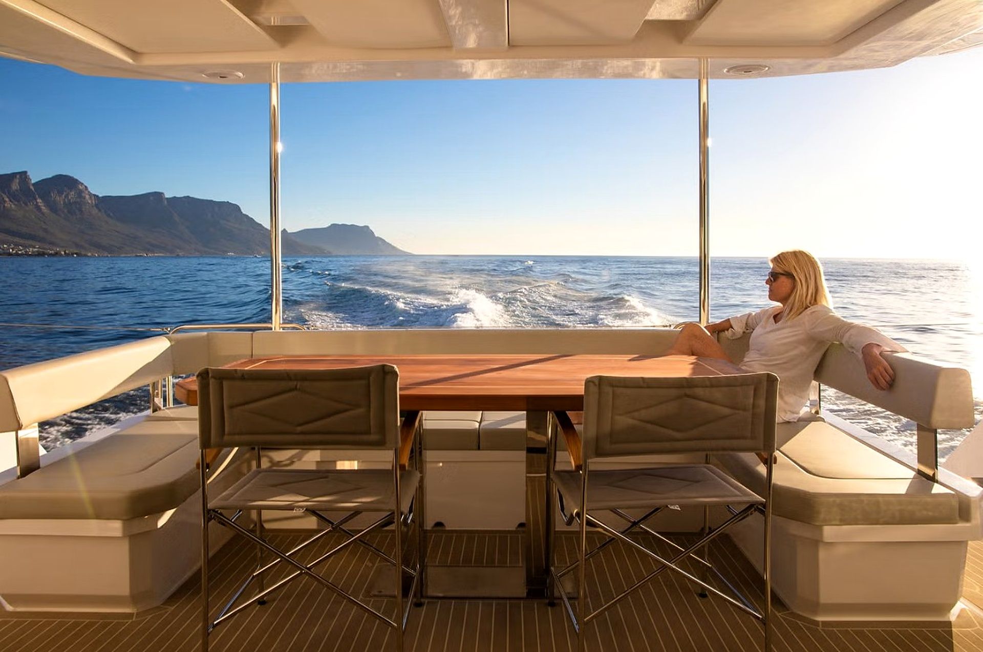 A boat interior with a table and chairs, large windows, and a view of the sea and mountains.