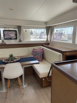 A compact interior with a dining area, cushioned seating, a small table with a decorative plant, and windows offering a marina view. - Sun-Kissed Sailing Adventure in Islas Sarónicas (Alimos)