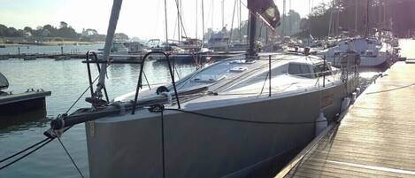 Un voilier est amarré dans une marina, avec un pont en bois et d’autres bateaux en arrière-plan.