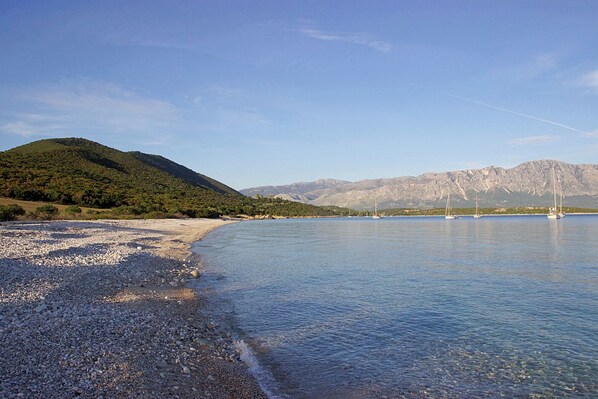 Une plage de galets avec des bateaux, une eau claire et des montagnes en arrière-plan.