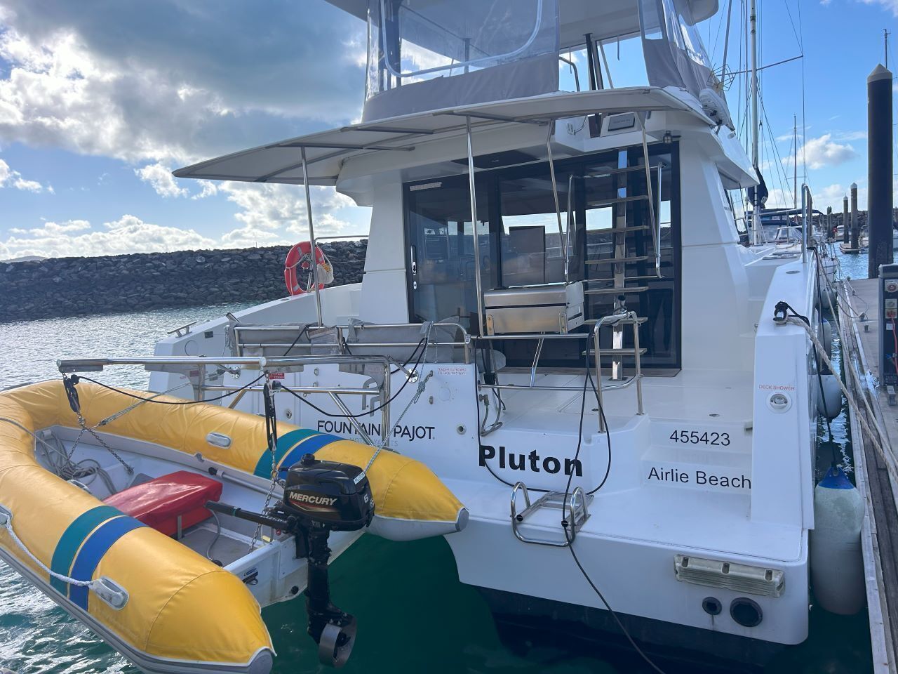 A boat named 'Pluton' docked at a marina with a lifebuoy, a deck shower, and a Mercury outboard motor.