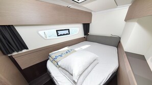 Quarto compacto e bem iluminado, com uma cama, uma pequena janela com vista e um painel de controle na parede.