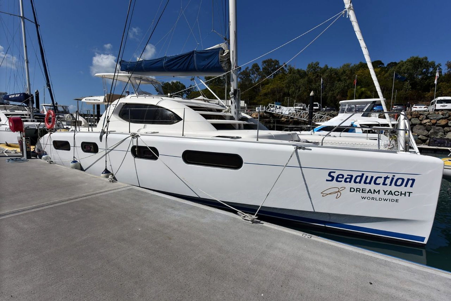 A white yacht named 'Seaduction' docked at a marina with other boats and a clear sky.