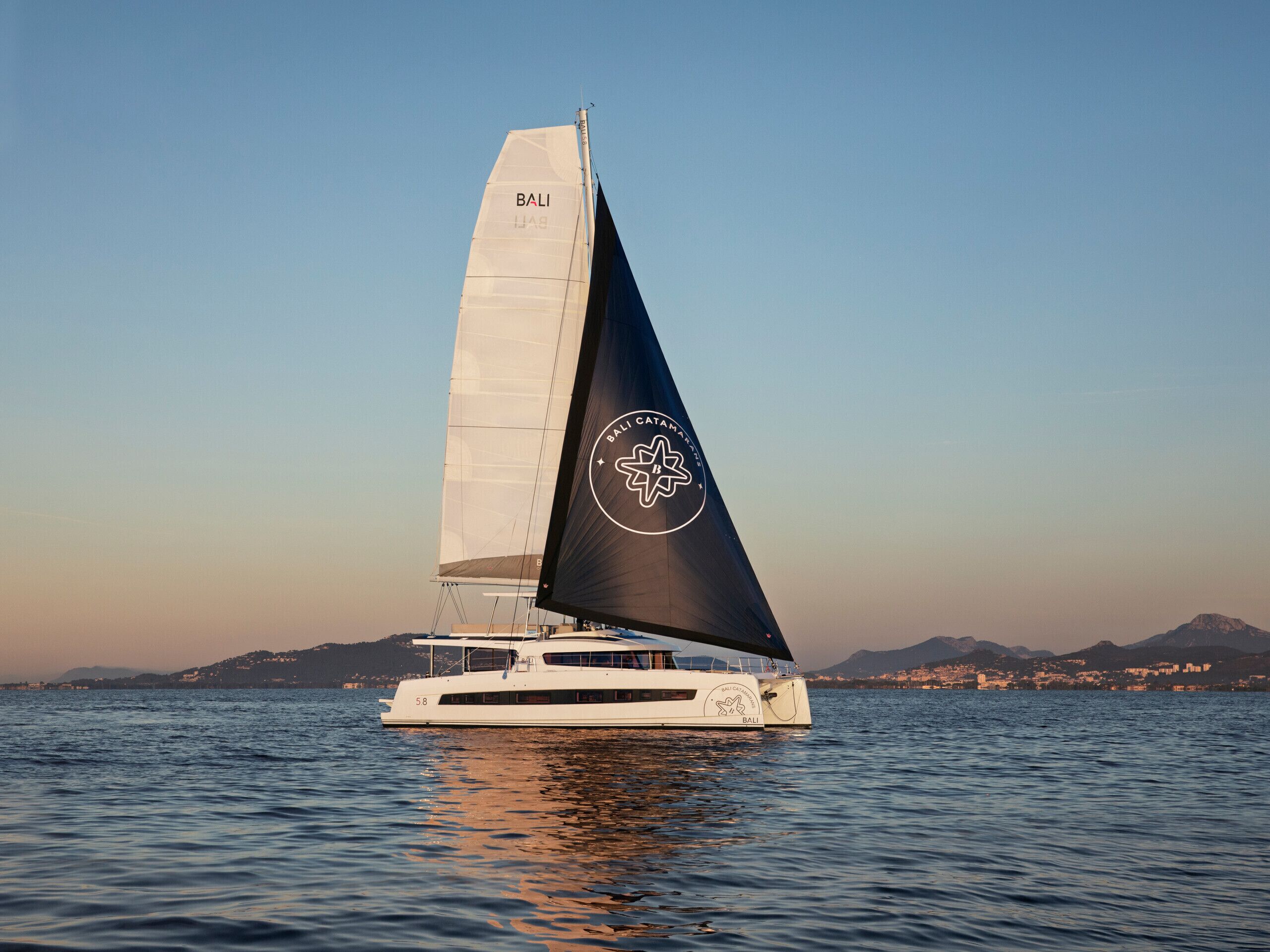 Un voilier arborant un logo sur sa voile et sa coque, naviguant sur une eau calme, avec une côte montagneuse en arrière-plan.