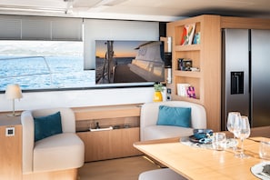 A modern interior with a TV displaying a scenic view, a dining area with a table set for two, and a kitchen area with a refrigerator.