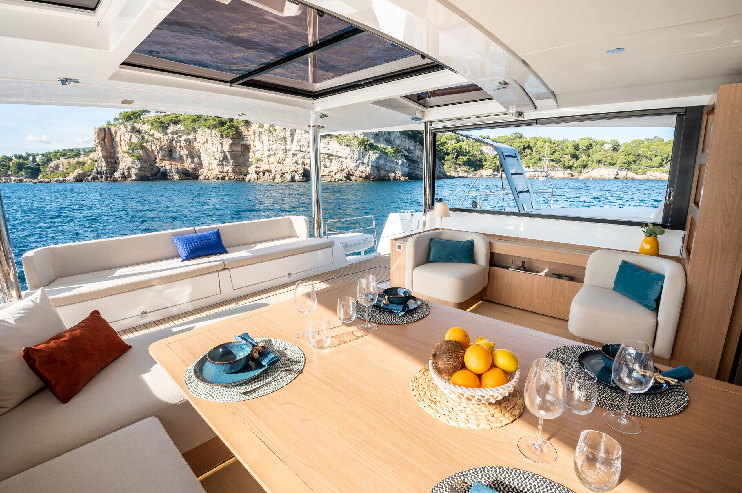 Interior de un barco con comedor, asientos y vistas al mar y acantilados.
